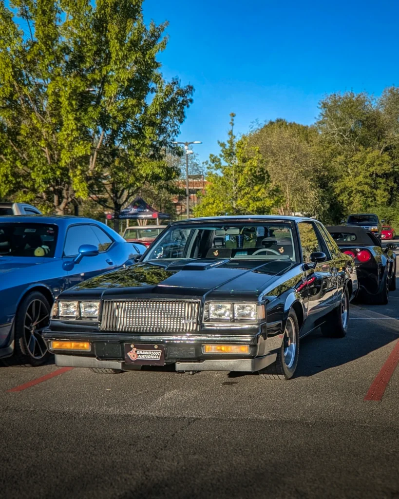 A black Buick Regal Grand National parked among other cars at an outdoor car show.