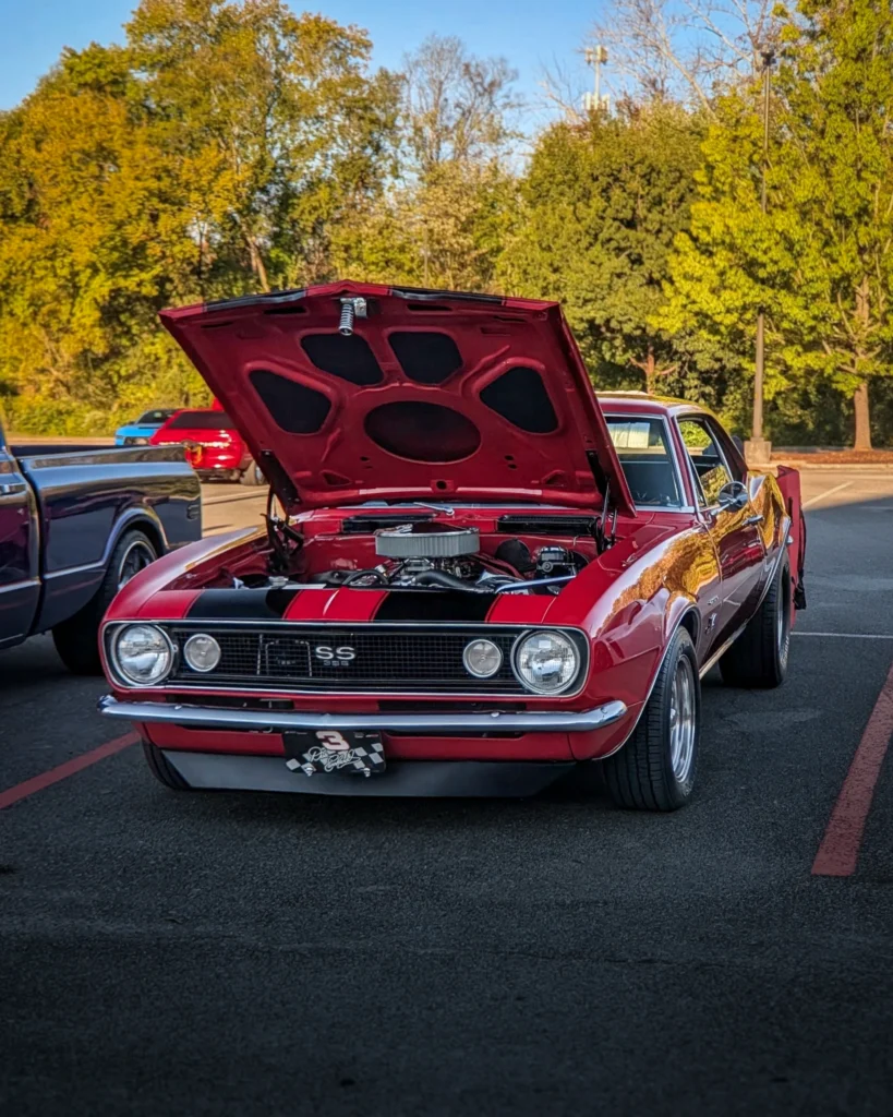 Red Chevrolet Camaro SS 396 with hood open showing engine at outdoor car event in parking lot with green trees in background