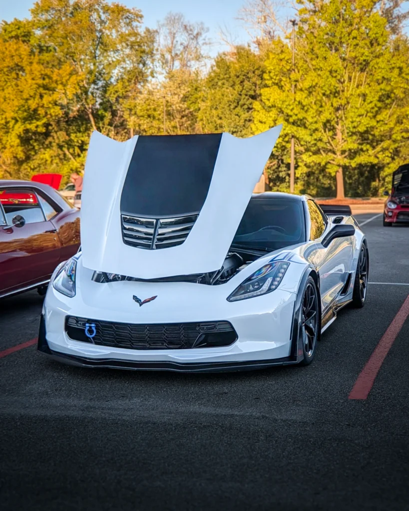 White Chevrolet Corvette with hood raised at outdoor car event with trees in background