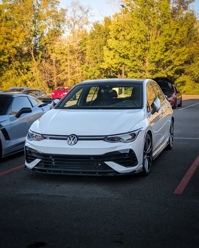 A white Volkswagen Golf R is parked among other cars in a tree-lined lot.