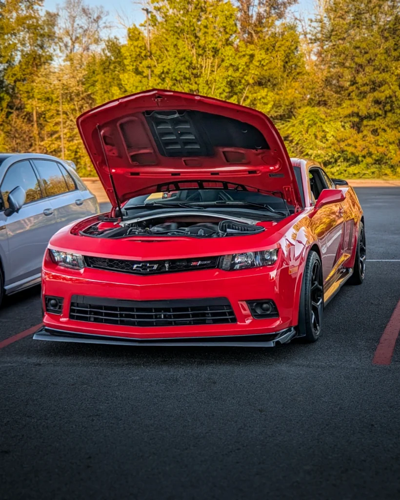 Red Chevrolet Camaro with hood open in parking lot with trees in background