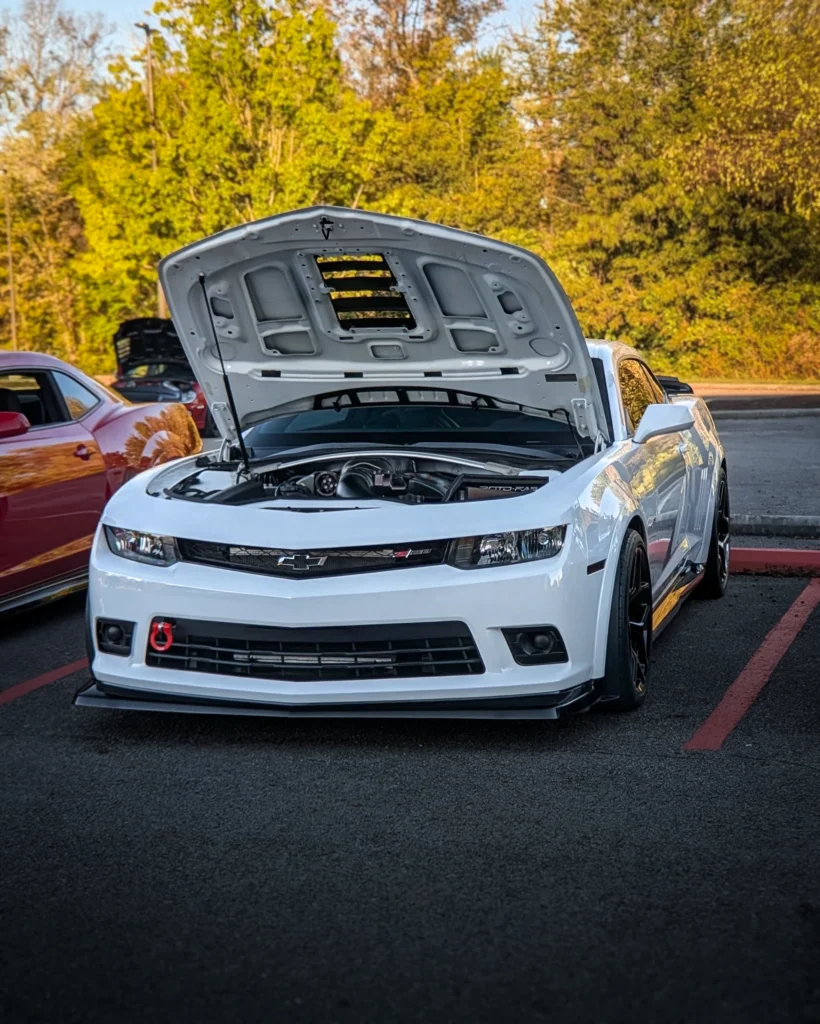 White Chevrolet Camaro with raised hood showing engine at outdoor car event with trees in background