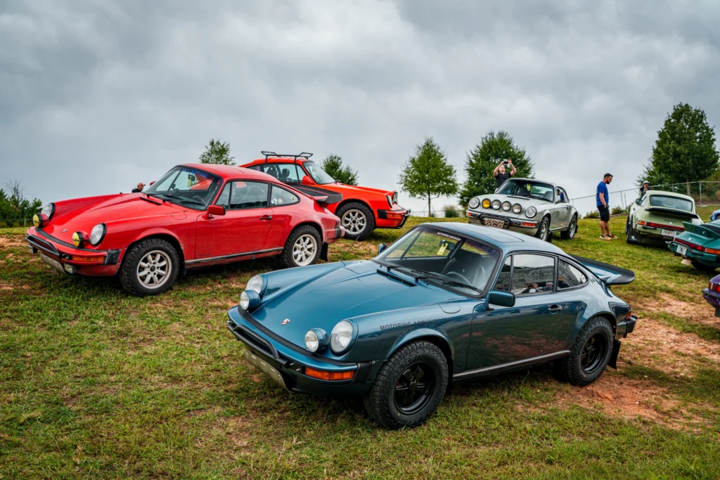 Five Porsche 911 cars in various colors parked on grass with trees and cloudy sky in background.