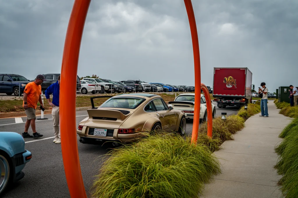 Two beige Porsche 911 sports cars parked on a road near a sidewalk with orange poles and people nearby under a cloudy sky