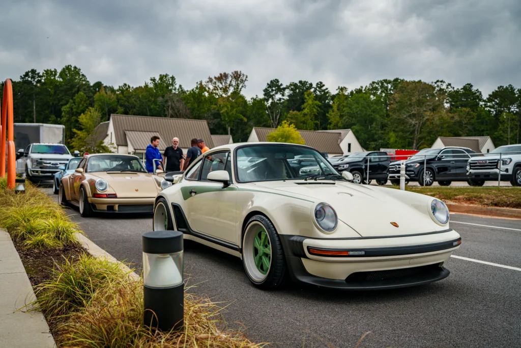 White Porsche 911 with green wheels parked in front of a beige Porsche 911 with people standing near the cars outside under