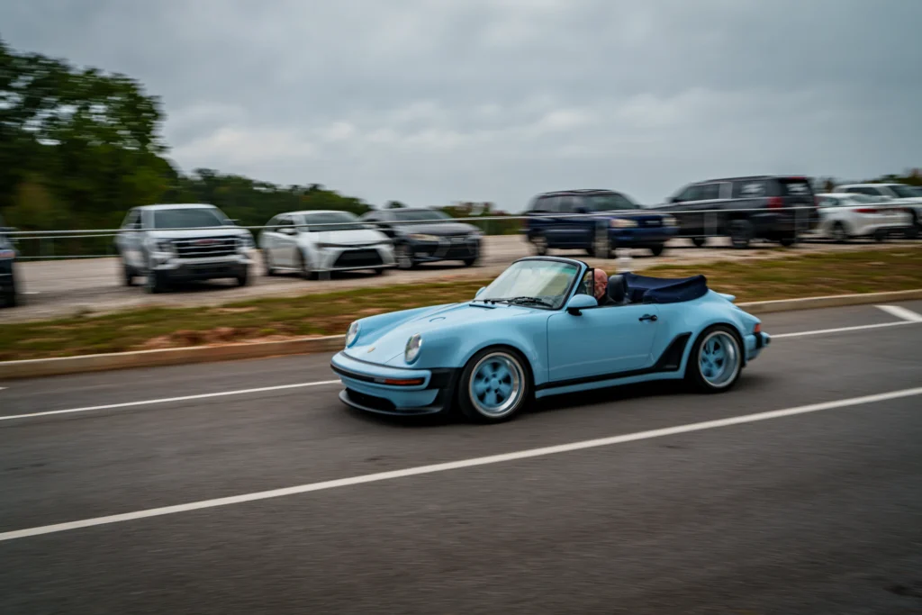 Light blue Porsche 911 convertible driving in parking lot with several parked cars under overcast sky