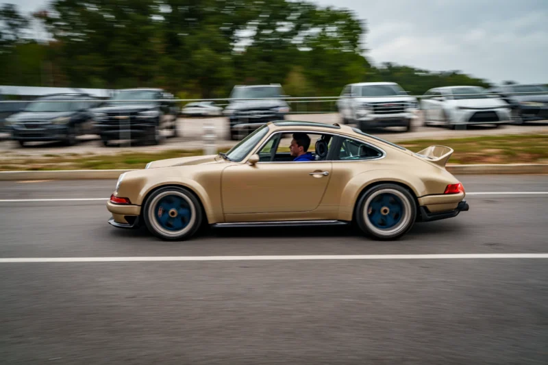 Tan Porsche 911 with blue wheels driving past a parking lot with SUVs and sedans in the background on a cloudy day