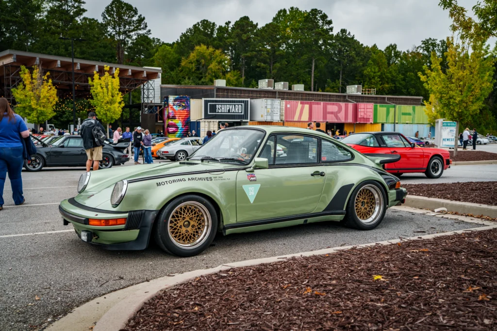 Light green Porsche 911 coupe parked at an outdoor car event with other vehicles and people in the background on a cloudy