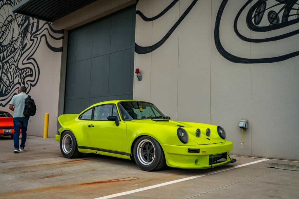 Bright green Porsche 911 parked beside a building with black abstract wall art and a man walking away with a backpack