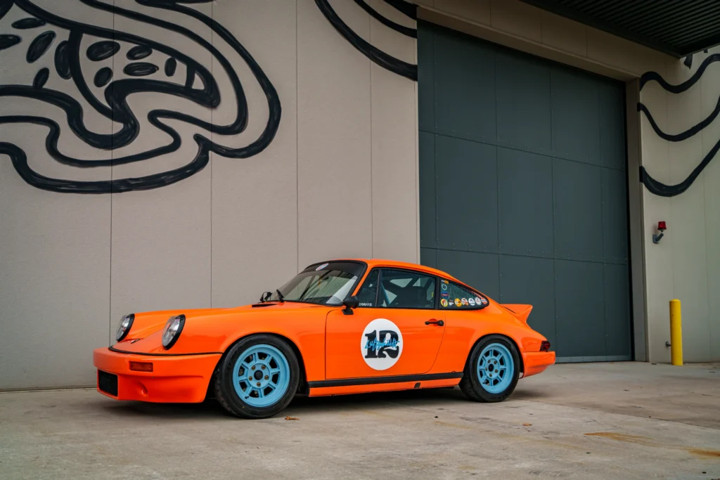 Orange Porsche 911 with blue wheels and racing decals parked by a building with graffiti art on the wall