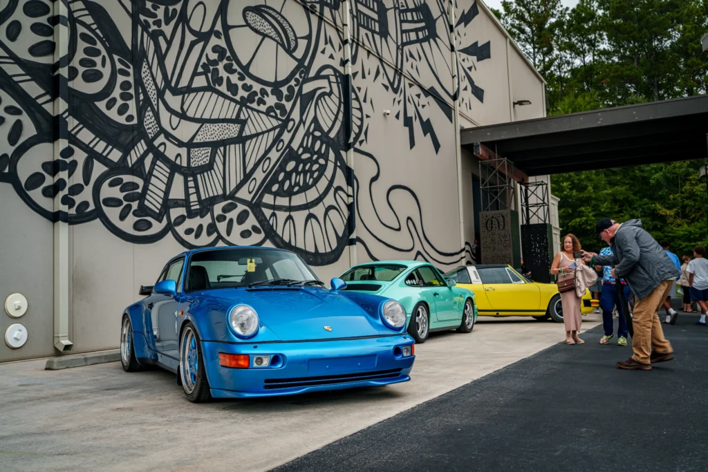 Blue Porsche 911 parked in front of a building with black-and-white mural, alongside green and yellow Porsche 911s; people