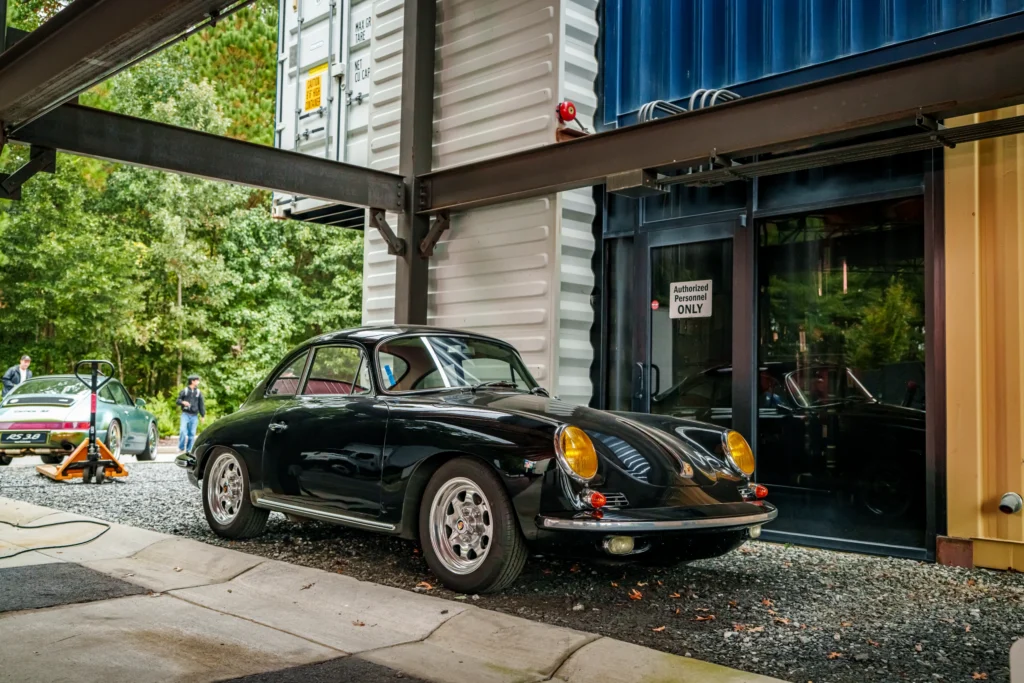 Black Porsche 356 coupe parked near shipping containers and glass door, green Porsche 911 RS 3.8 in background outside near