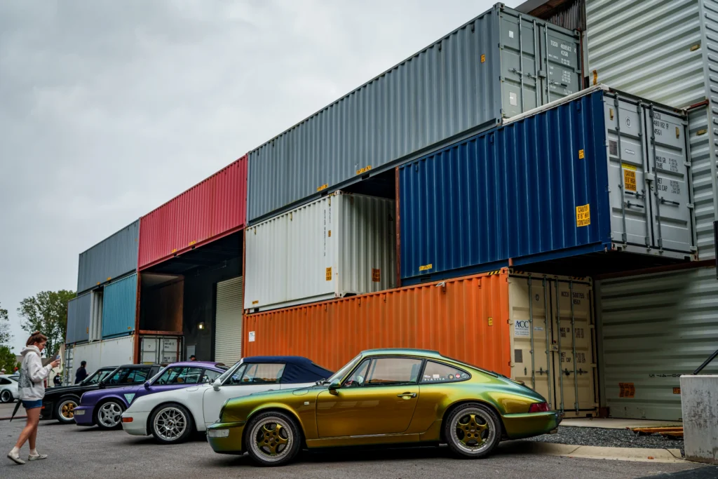 Green Porsche 911 parked near stacked shipping containers with other cars and people nearby at a car event.