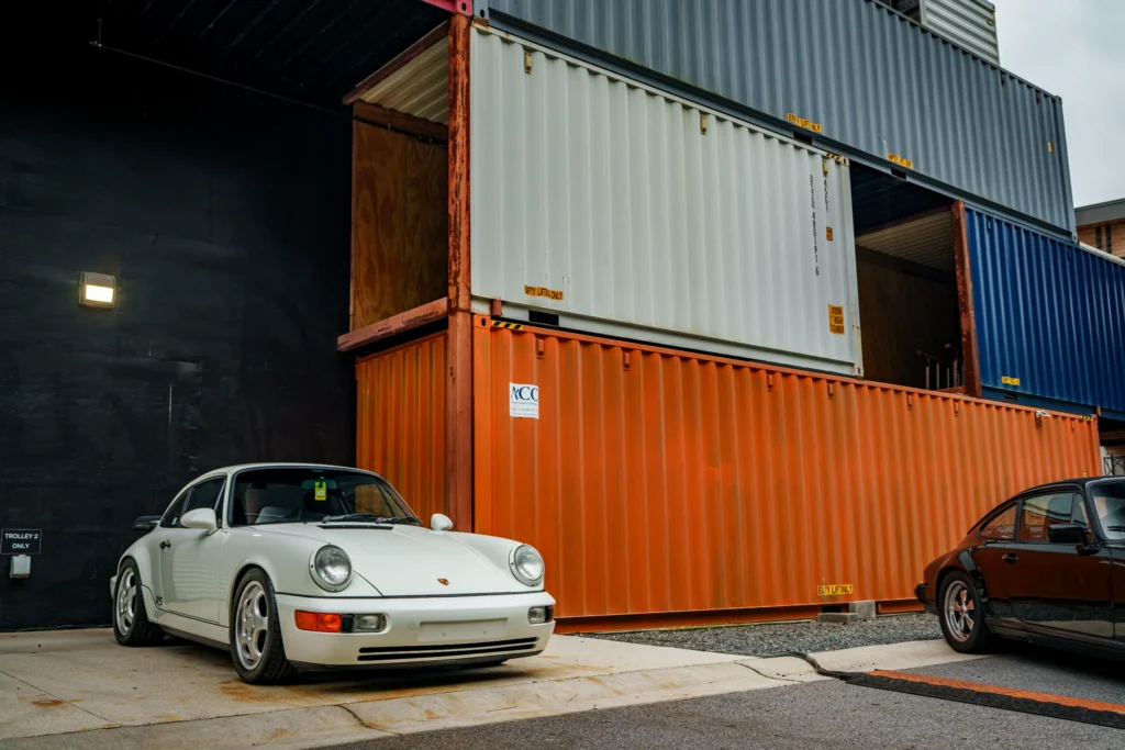 White Porsche 911 RS parked near stacked shipping containers in an industrial setting