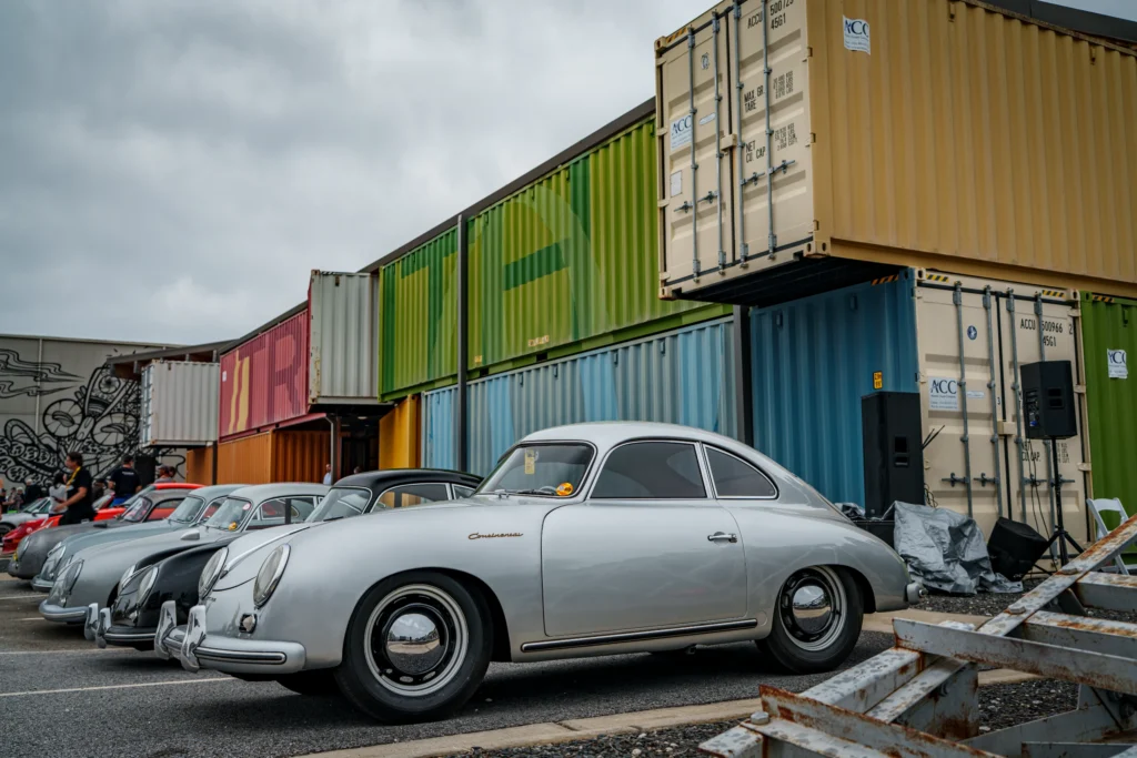 Silver Porsche 356 Continental parked in front of stacked colorful shipping containers at an outdoor car event.