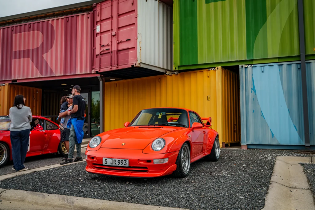 Red Porsche 911 parked in front of stacked colorful shipping containers with people standing nearby