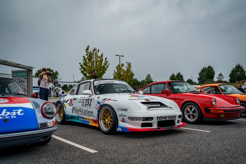 White Porsche 911 GT1 race car and red Porsche 911 coupe parked outdoors with people and trees in the background