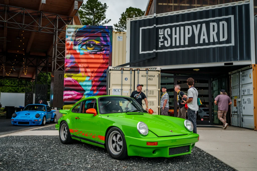 Bright green Porsche 911 Carrera parked outside The Shipyard with a blue Porsche in the background and several people nearby.