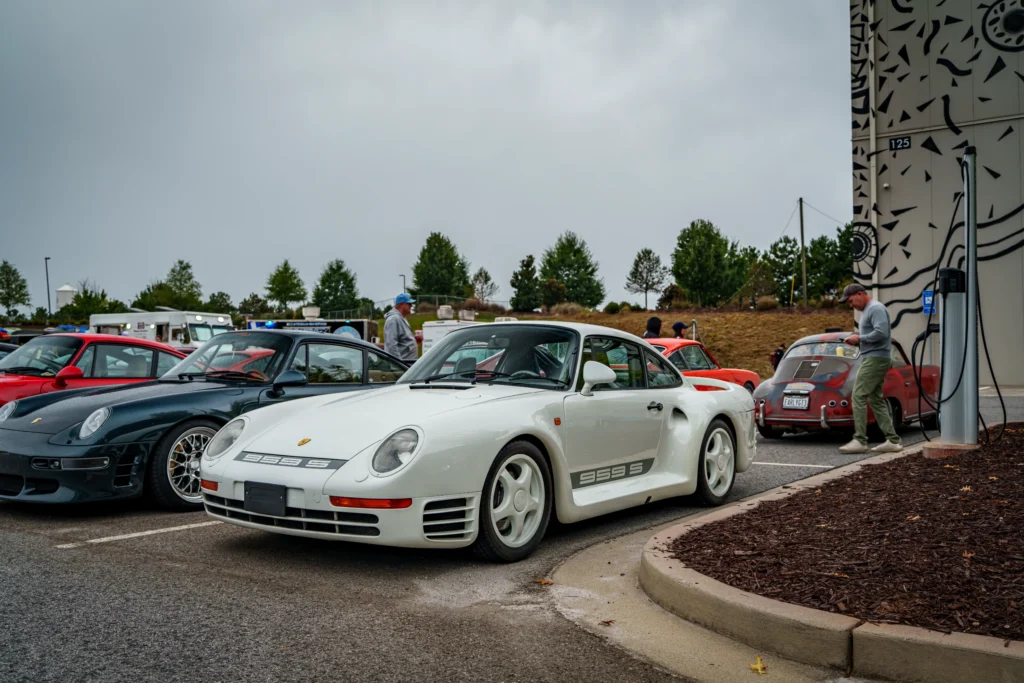 White Porsche 959 S parked in a row with other Porsche cars in an outdoor parking lot with people and trees nearby