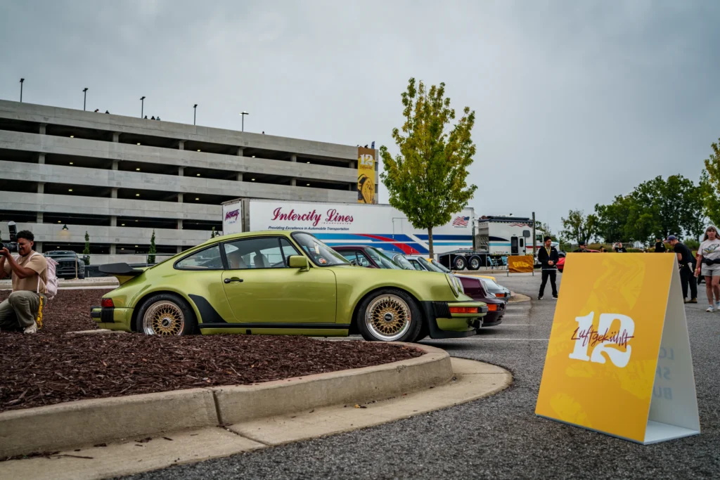 Green Porsche 911 parked in a lot next to a crowd and event signage under an overcast sky
