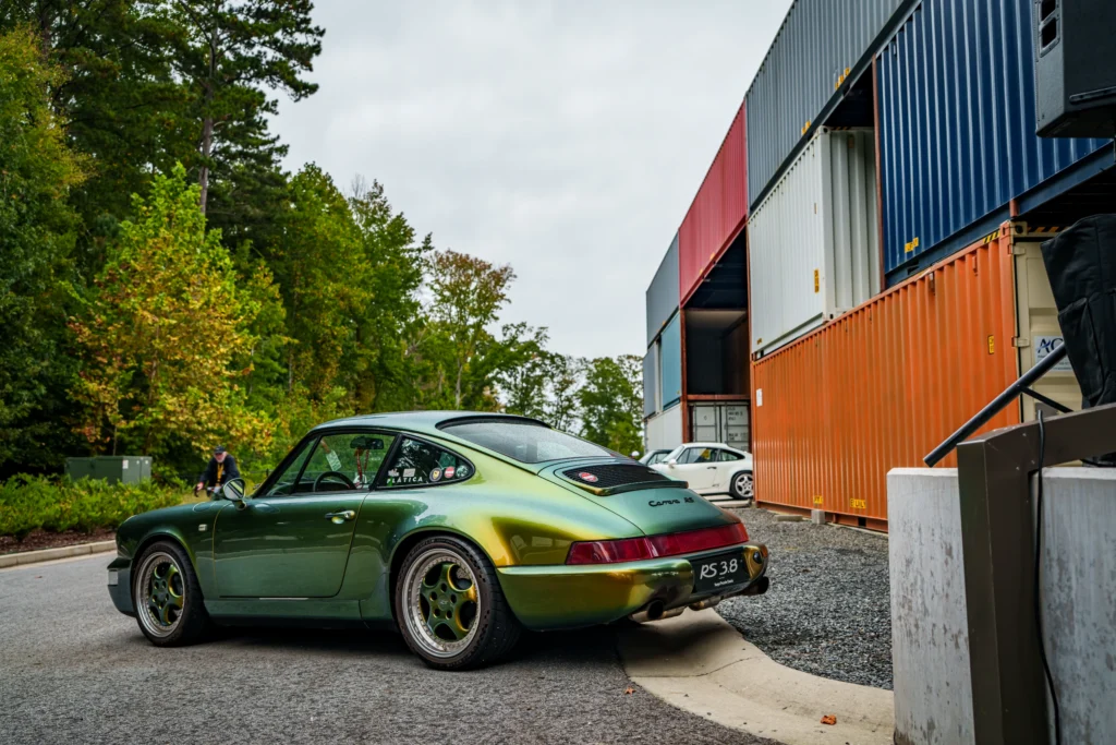 Green Porsche 911 Carrera RS 3.8 parked beside stacked shipping containers with guy on bicycle in background
