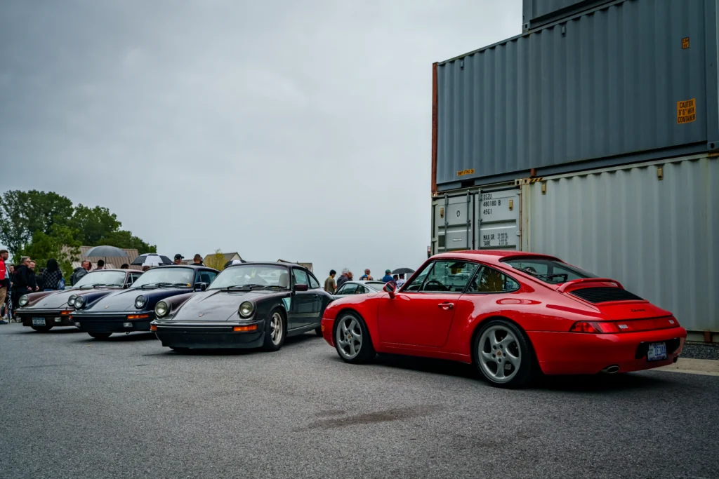 Four Porsche 911 coupes in various colors parked in a row outdoors with a crowd and container in background