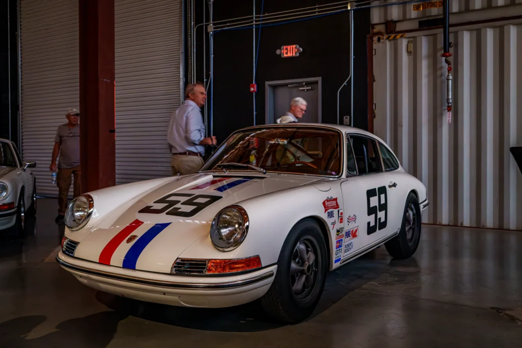 White Porsche 911 with red and blue racing stripes and number 59 parked indoors with people walking nearby