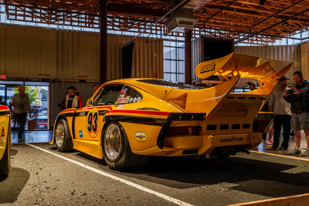 A yellow Porsche 935 race car with number 93 and a large rear wing sits indoors among attendees.