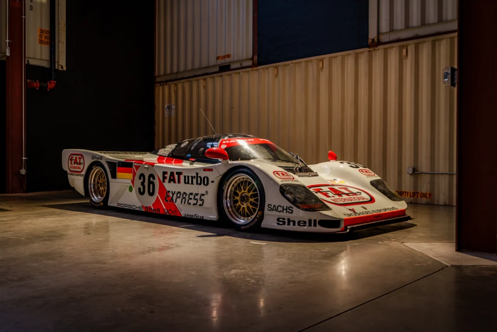 White Porsche 962 race car with red and black decals inside a warehouse with stacked shipping containers