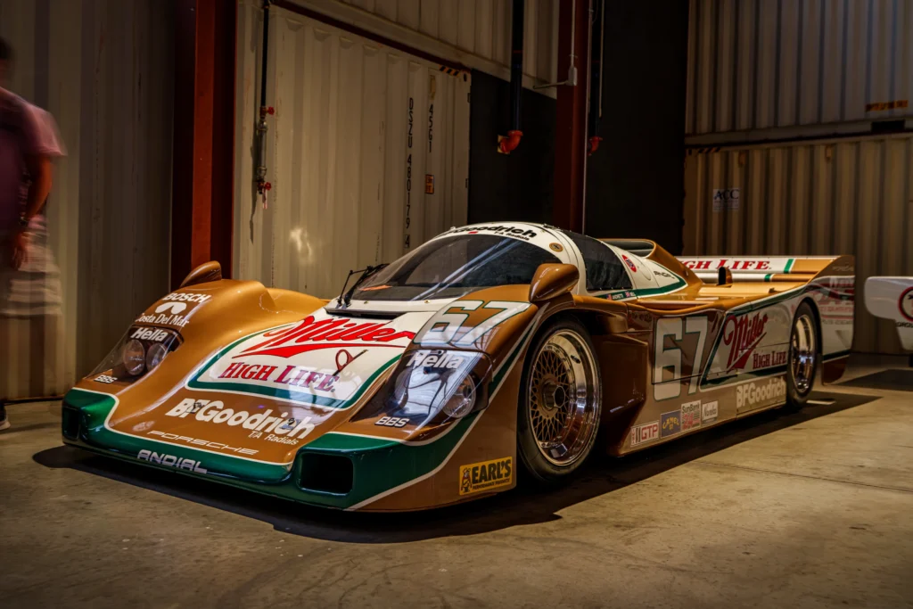 White and gold Porsche 962 race car inside a storage or garage area with numbered shipping containers in the background