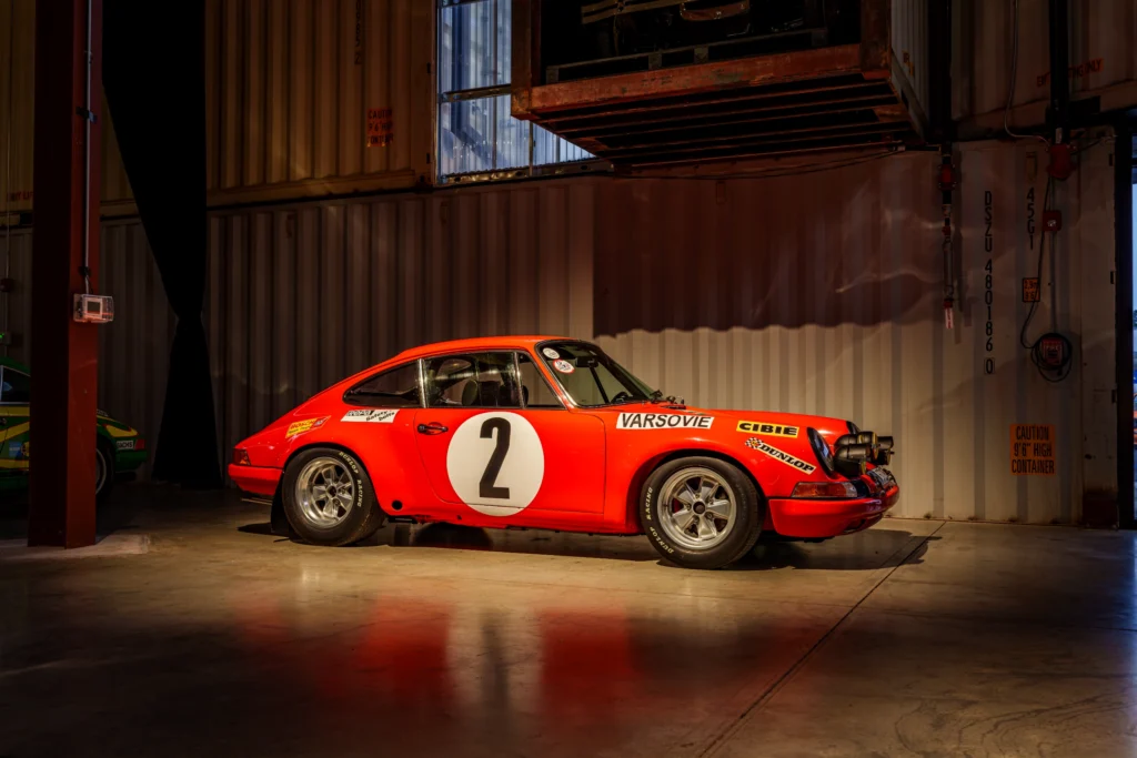 Red Porsche 911 with racing decals and number 2 on a white circle, photographed indoors from the driver's side under