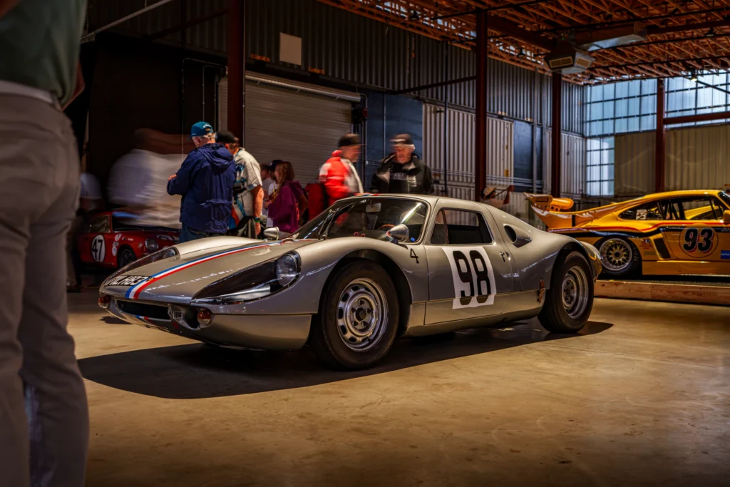 Silver Porsche 904 race car with number 98 and a yellow Porsche 935 race car inside a garage with people around.