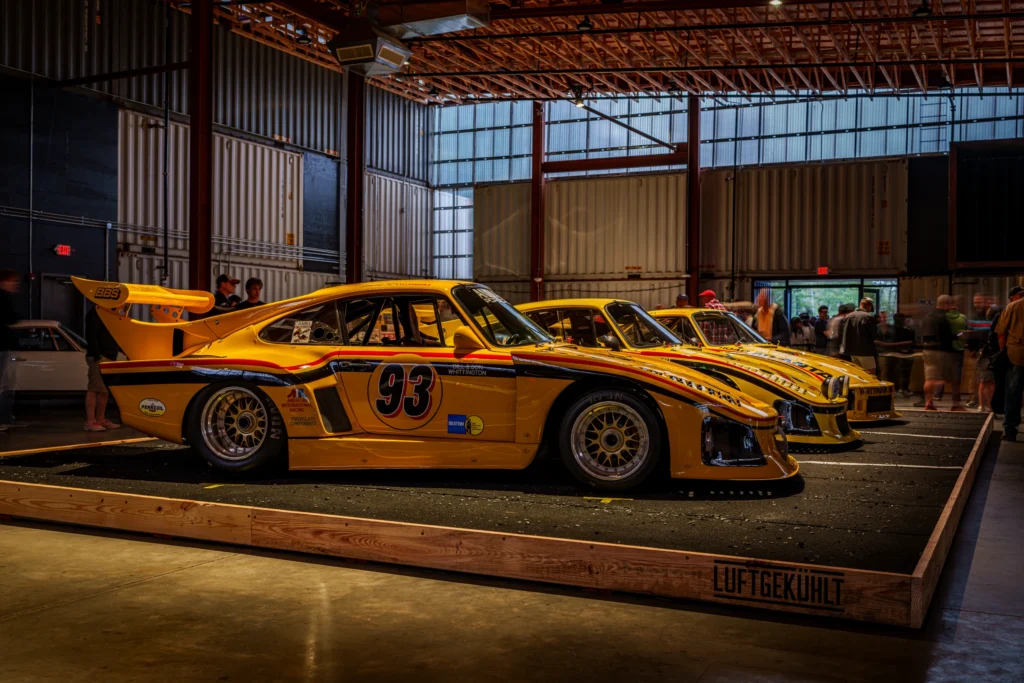 Yellow race cars in a row inside a large industrial building with people walking nearby