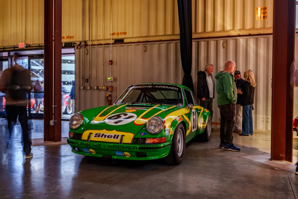 Green Porsche 911 race car with Shell and Kremer livery inside an event hall with people standing nearby