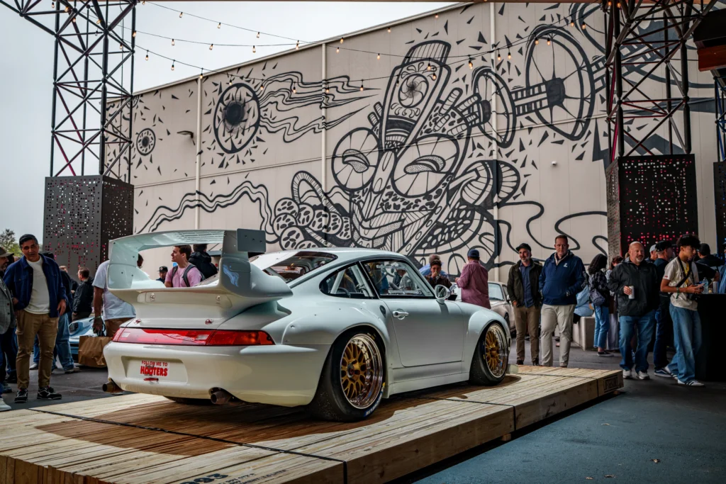 White Porsche 911 with large rear wing on wooden platform with people and a mural in the background.