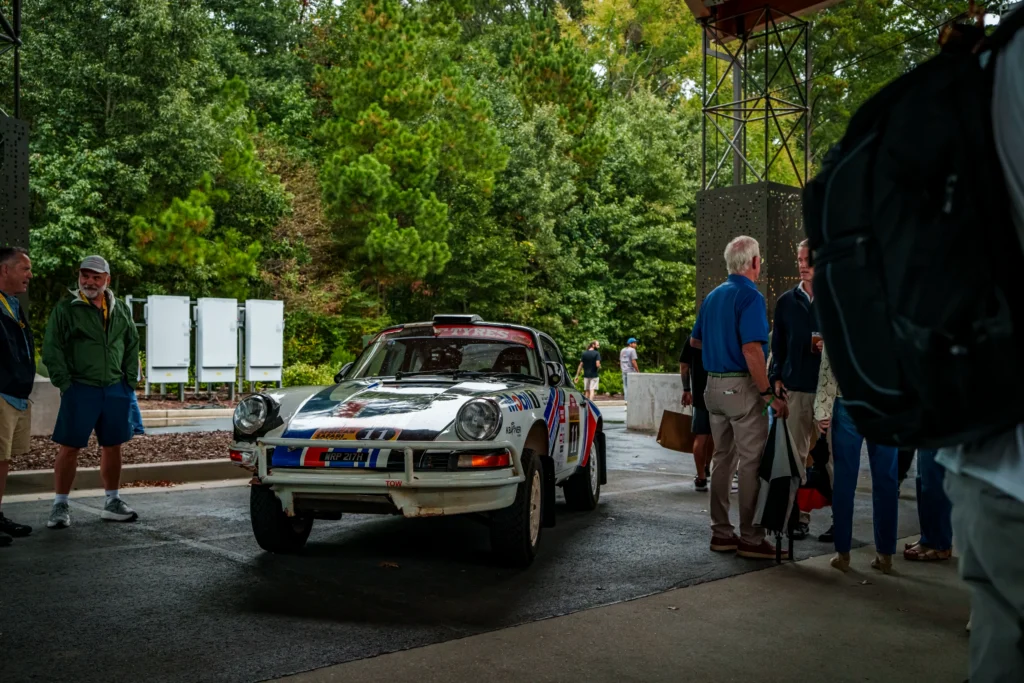 White Porsche 911 rally car with decals parked under a canopy with people nearby and trees in the background