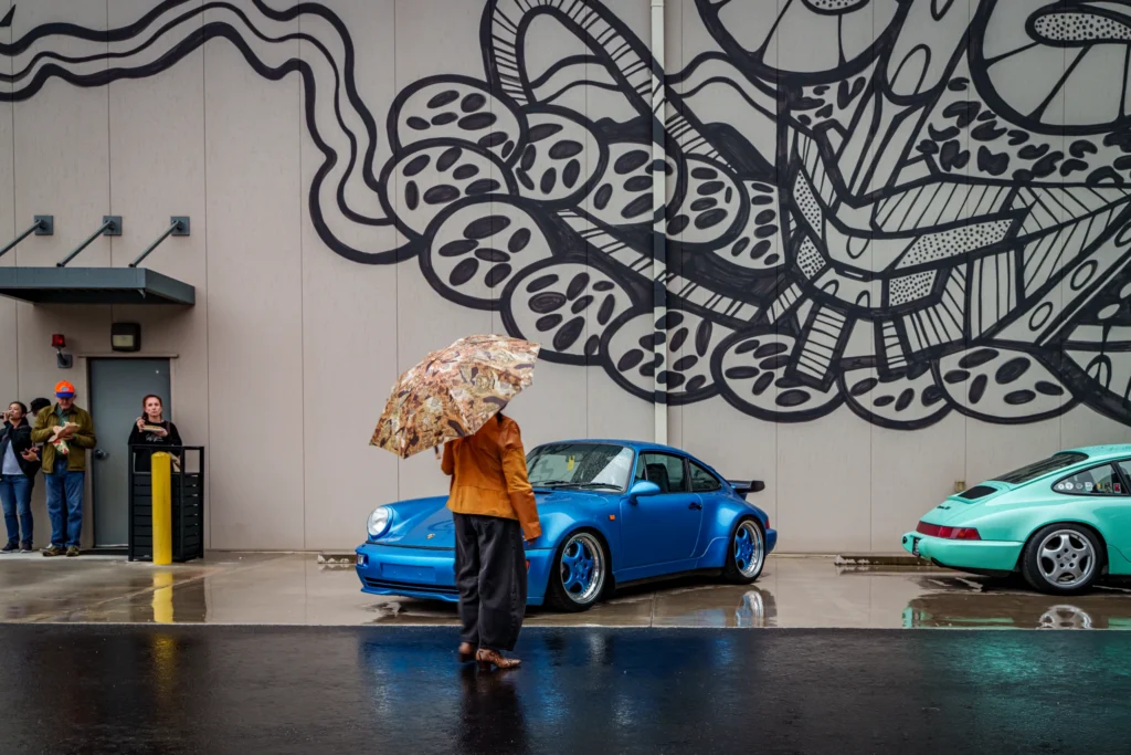 Blue Porsche 911 parked in front of a wall mural with people nearby and a person holding a patterned umbrella