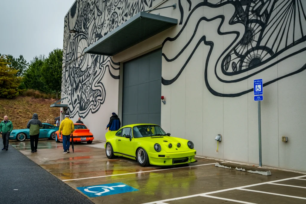Three Porsche 911 cars in green, orange, and teal parked beside a building with black-and-white mural art and people walking