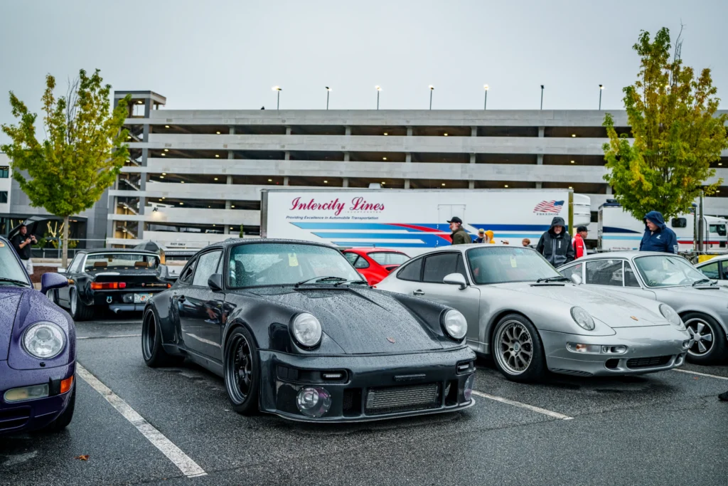 Black Porsche 911 parked next to a silver Porsche 911 with rain droplets visible on the cars in an outdoor parking lot near