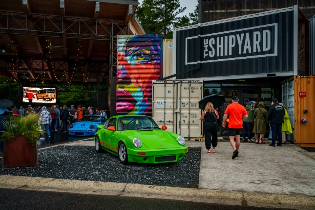 Green Porsche 911 and light blue Porsche 911 parked outside a venue with a crowd entering nearby
