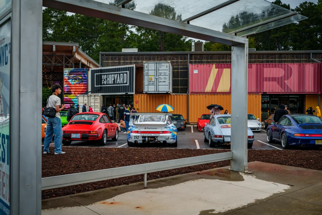 Parking lot with multiple Porsche vehicles in various colors near a building made from shipping containers with people around