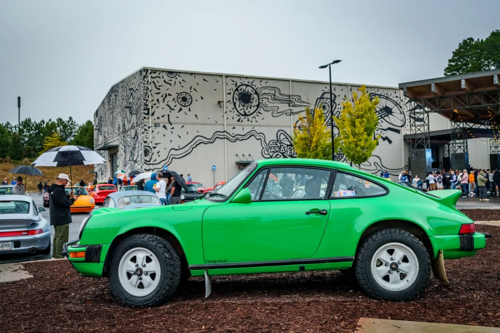 Green Porsche 911 with off-road tires parked outdoors near a building with mural, surrounded by people with umbrellas at a