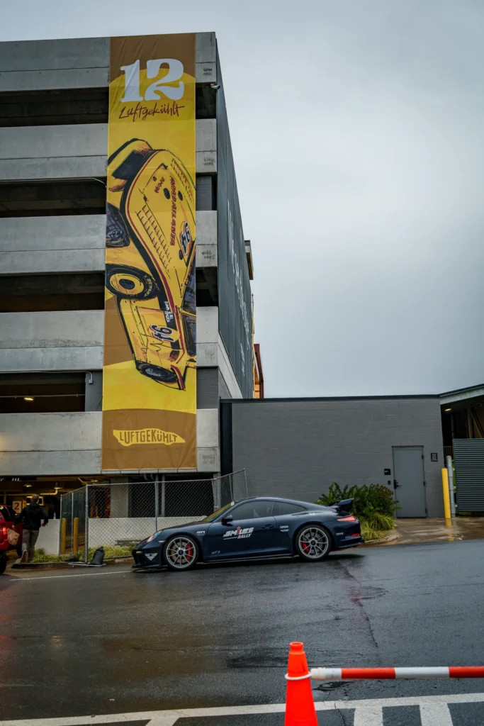Blue Porsche 911 GT3 parked by a gray building with a large yellow banner featuring a vintage race car illustration