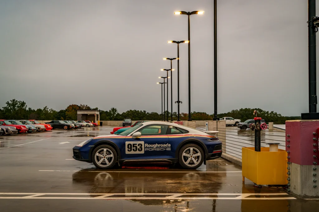 Blue and white Porsche 911 with number 953 decal parked on wet rooftop under lamp posts and cloudy sky