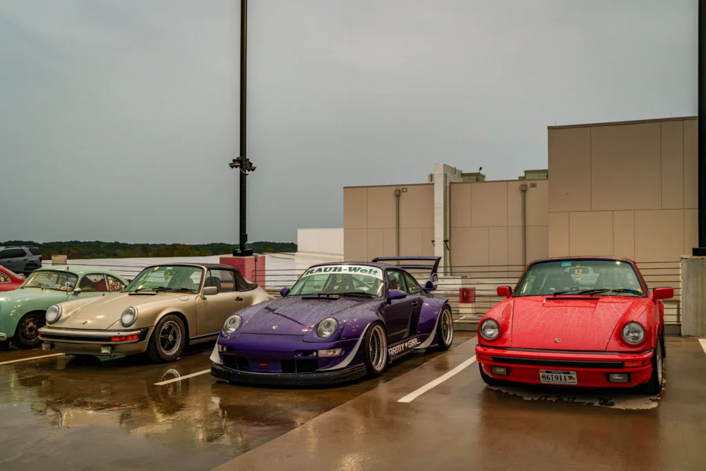 Purple, red, and beige Porsche 911s sit beside a light green Porsche 356 on a wet rooftop parking deck.