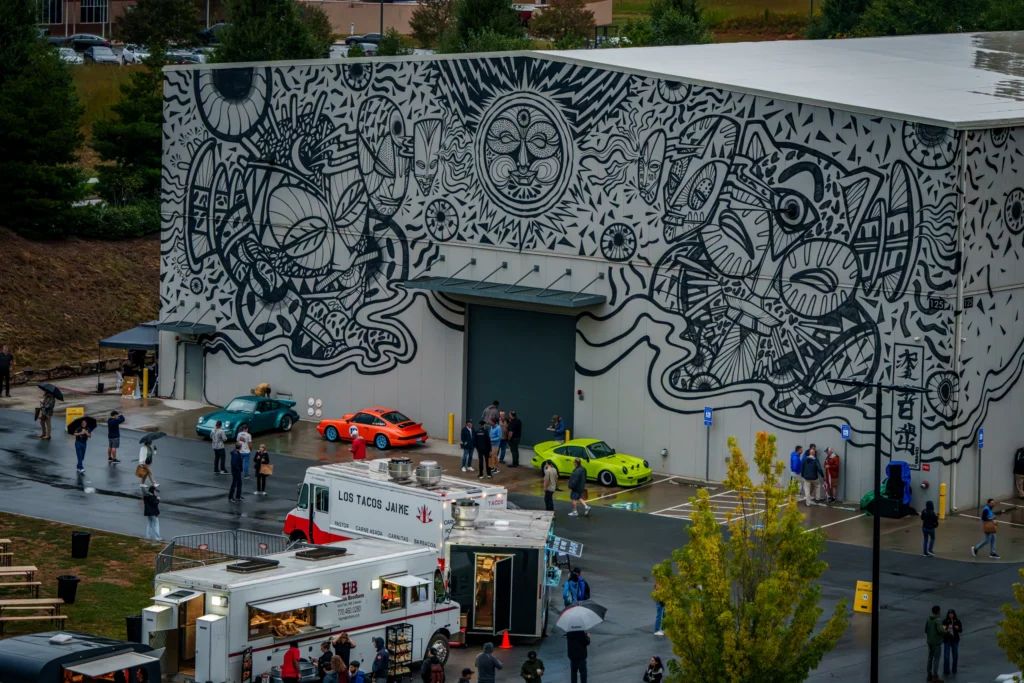 Three Porsche 911 cars in green, blue, and orange parked in front of a building with black and white mural and people nearby