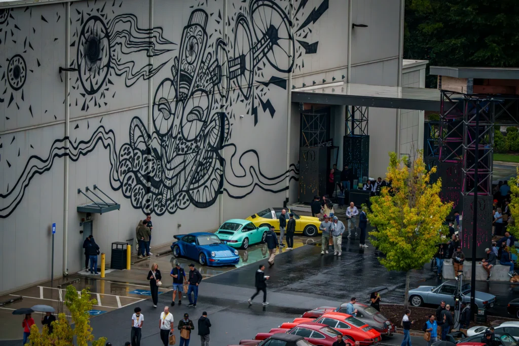 Yellow, turquoise, and blue Porsche 911s parked near a large building with black abstract mural, surrounded by walking