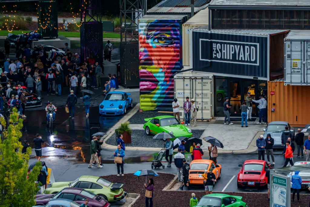 Crowd browsing cars at an outdoor automotive event with various Porsche models and colorful building murals