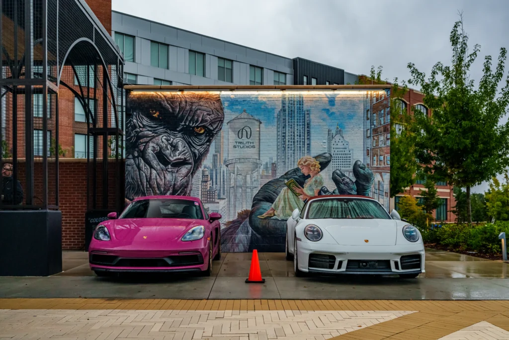 A pink Porsche Boxster and a white Porsche 911 sit on wet pavement in front of a King Kong mural.