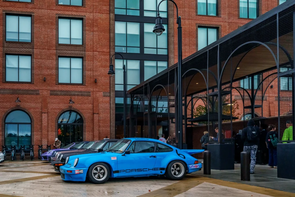 Blue Porsche 911 parked in front of a brick building with other cars and people nearby during an outdoor event.